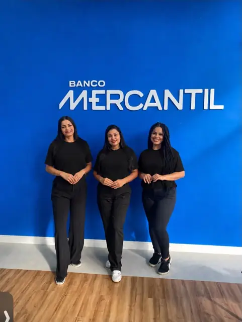 Três promotoras de preto em frente ao painel do Banco Mercantil